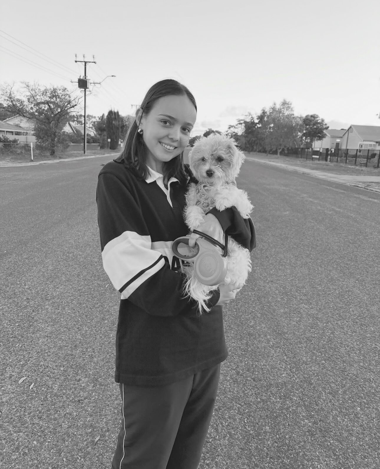 Adolescente sosteniendo un perro blanco esponjoso y parada en medio de una calle desierta, imagen en blanco y negro. 