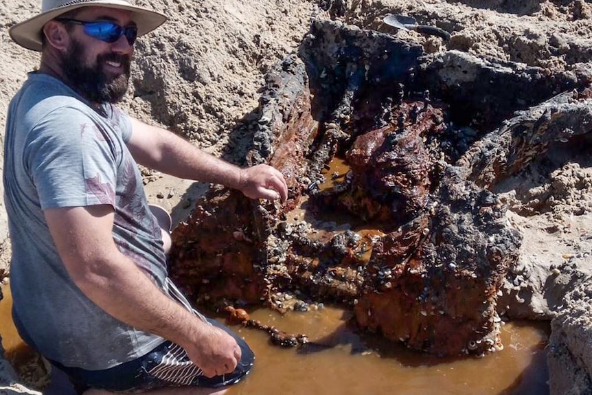 A man in a cowboy hat with a rusty car body buried in the sand.