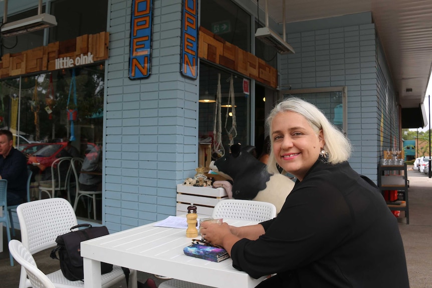 A blonde woman sits as a cafe table, smiling.