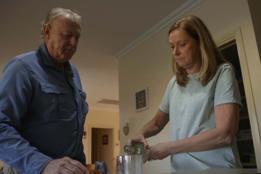 A man and a woman stand beside a kitchen bench. The woman pours water out of a glass bottle into a cup