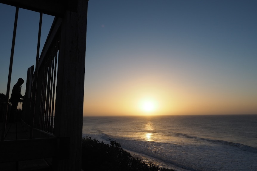 The silouette of a man standing on a timber lookout, looking at the ocean, surfers and the sunrise.