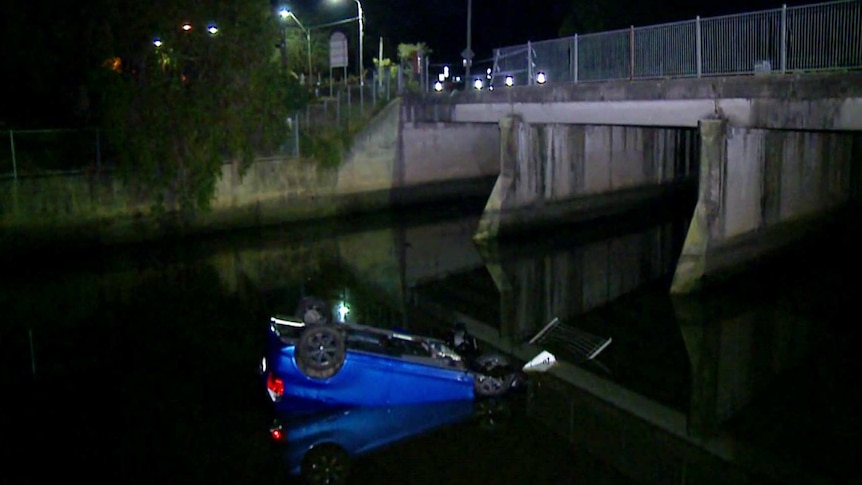 A blue car is submerged on its roof in a river, with police tape set up across where a bridge railing is missing.