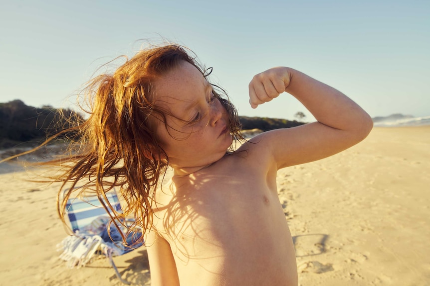 A child at the beach with shoulder-length hair flexes one arm's bicep while looking towards the muscle.