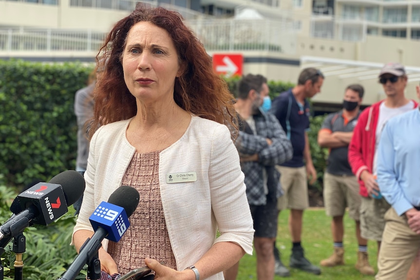 A woman stands in front of a group of tradesman with a cream jacket, talking to media in front of microphones