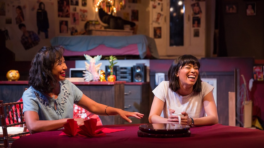 Colour production still of actors Hsiao-Ling Tang and Alex Lee seated and laughing at dinner table on stage.