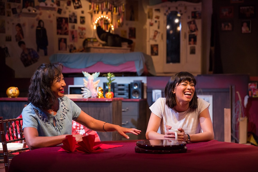 Colour production still of actors Hsiao-Ling Tang and Alex Lee seated and laughing at dinner table on stage.