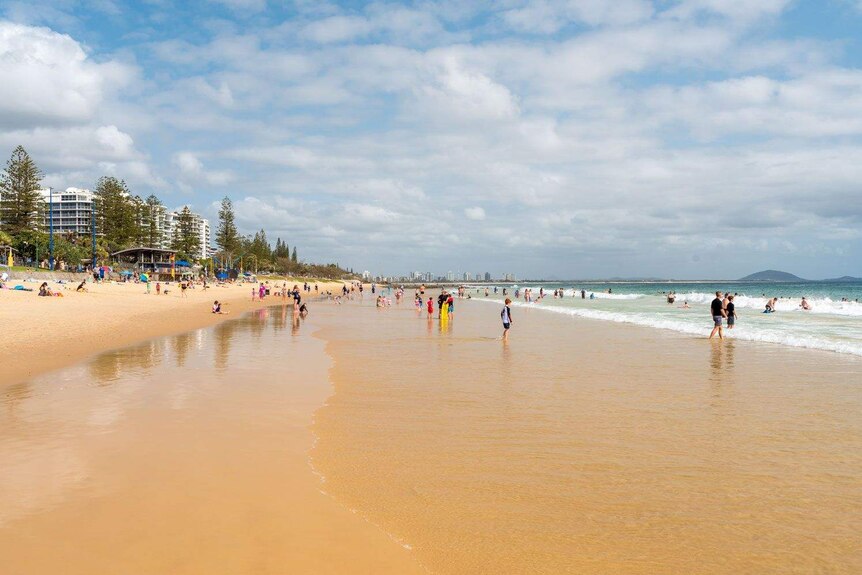 A wide shot of Mooloolaba Beach with people dotted across the sand and in the water.