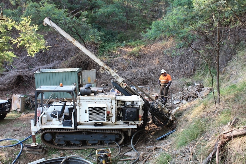 A man in a bright orange shirt looks at a machine that is drilling into the earth.