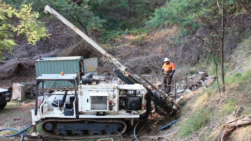 A man in a bright orange shirt looks at a machine that is drilling into the earth.