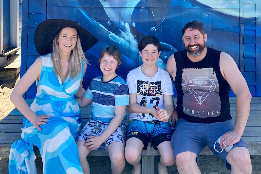 A woman, man and two young children smile, dressed in blue and sitting in front of a blue dolphin mural at the beach.