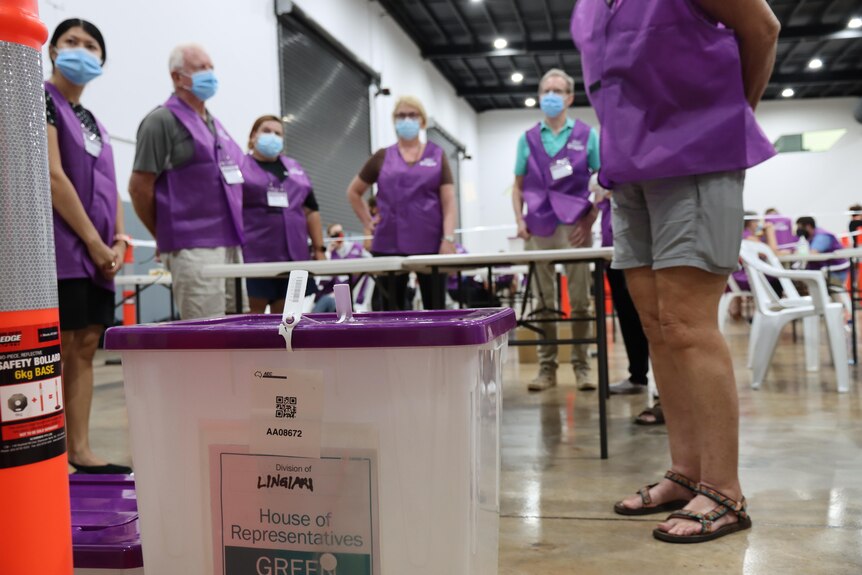 Feet can be seen of people in purple uniforms wearing face masks. There are ballot boxes nearby.