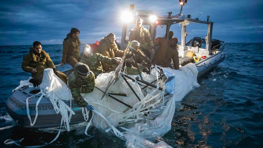 A group of men in military gear on a small military boat in the evening, pulling a large parachute or balloon up from the water