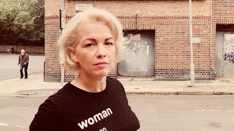 A woman with short blonde hair poses for a photo on the street wearing a black t-shirt that says 'woman' on it