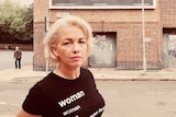 A woman with short blonde hair poses for a photo on the street wearing a black t-shirt that says 'woman' on it
