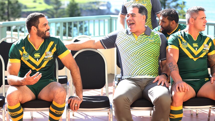 Two men sit on chairs laughing, the man on the left in full rugby league kit.
