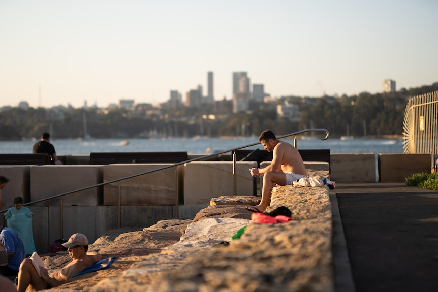 a man wearing swimmers sits on the side of a harbour pool in sydney while looking at this phone