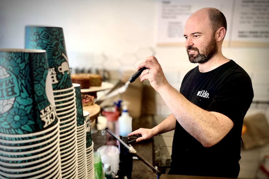 A balding man wearing a black tshirt slices cake in his cafe with blue coffee cups in the foreground