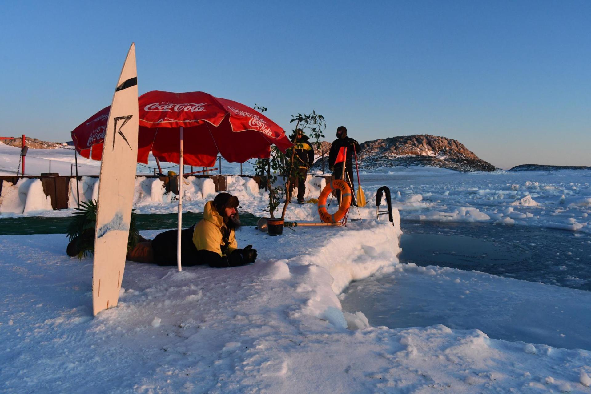 A man in full Antarctic gear lies on the snow, near an icy swimming hole, with a surf board and umbrella 