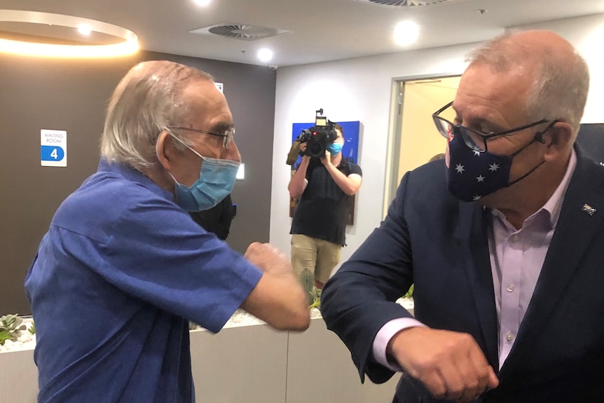 An elderly man wearing a medical face mask touches elbows with Prime Minister Scott Morrison who is also wearing a face mask. 