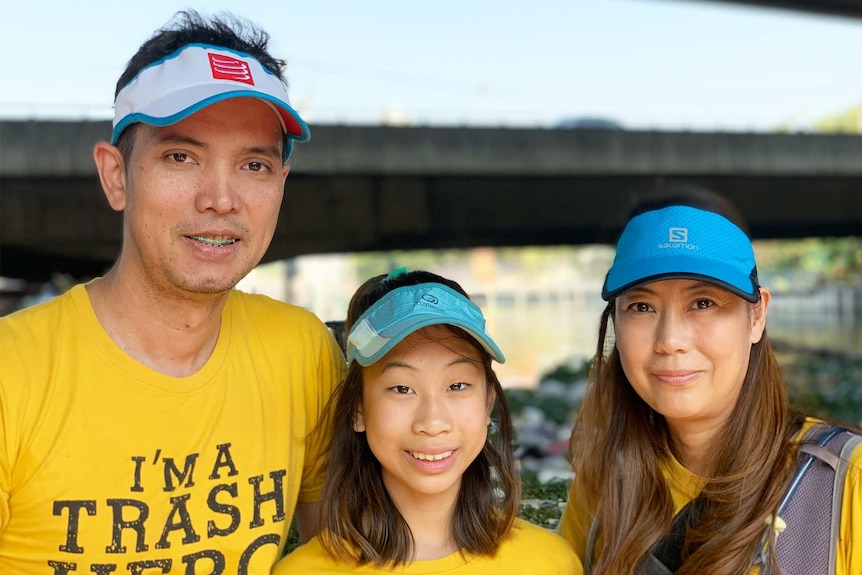 A man, girl and woman stand in front of a canal full of rubbish.