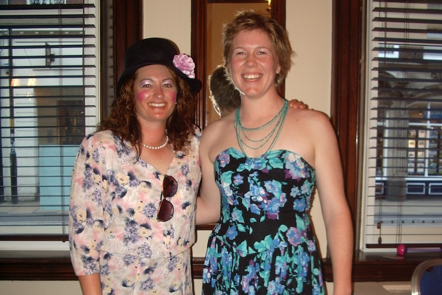 Two women in fancy dress pose and smile for the camera. They are wearing floral dresses, and Karen Rolton has a hat on.
