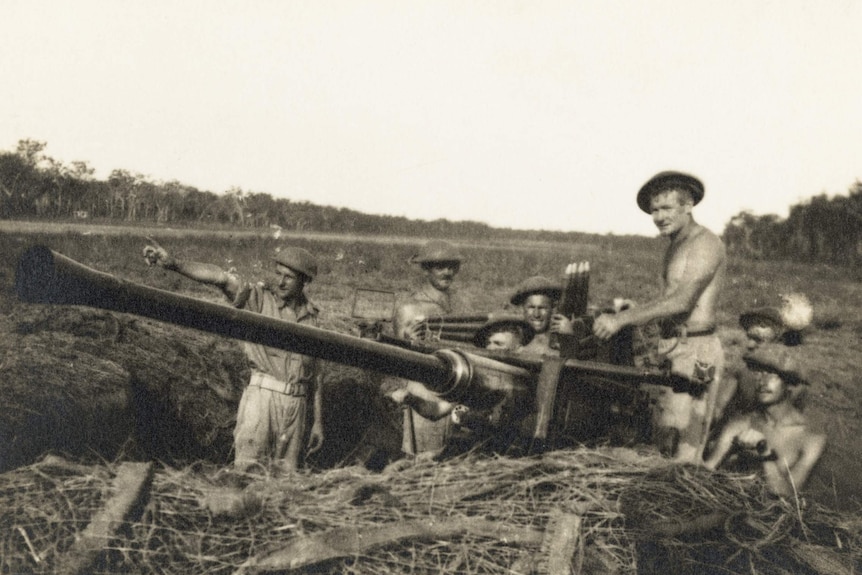 A historical black and white photo showing a group of men around a WWII-era cannon gun, in a field. 