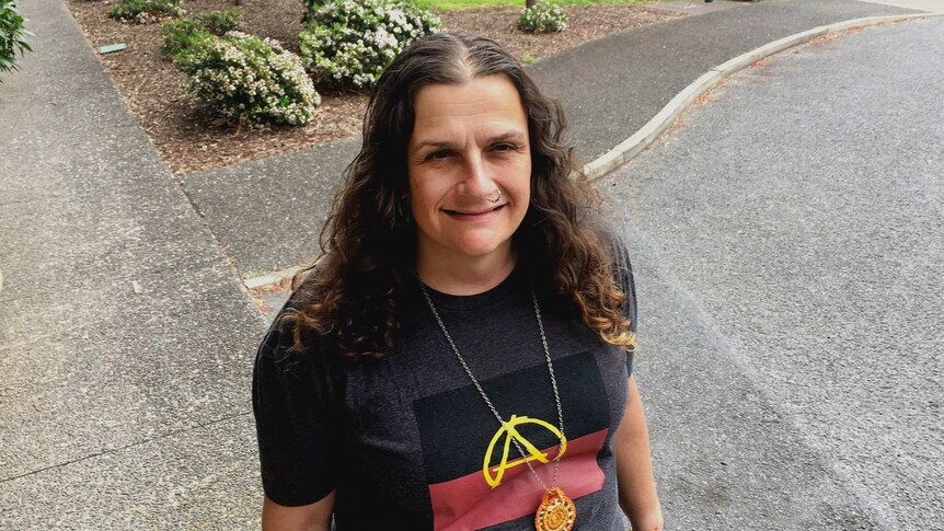 A woman stands outside wearing a t-shirt with the Aboriginal flag and anarchy symbols