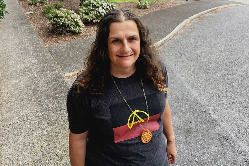 A woman stands outside wearing a t-shirt with the Aboriginal flag and anarchy symbols