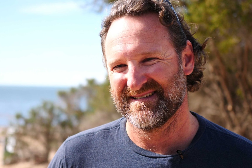 A man with a beard smiles at the camera. Water and trees can be seen in the background.