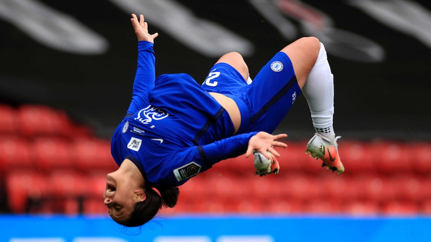 Sam Kerr, wearing blue football kit, appears in mid-flip, with her legs bent, arms by her side, upside down