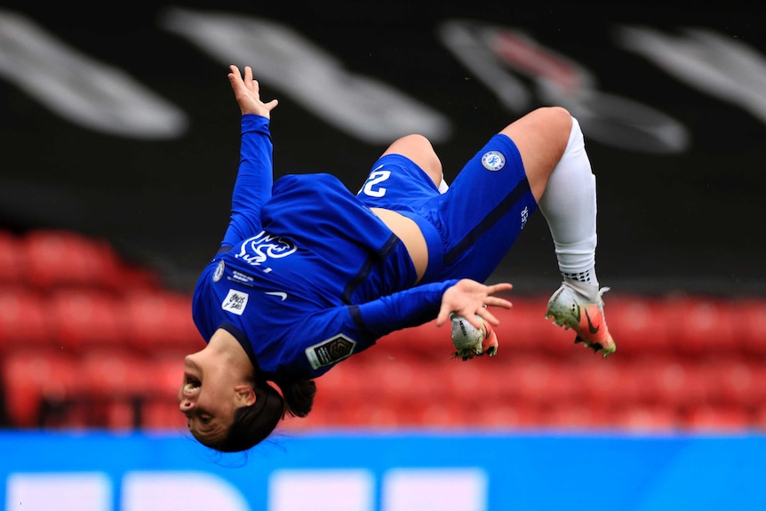 Sam Kerr, wearing blue football kit, appears in mid-flip, with her legs bent, arms by her side, upside down