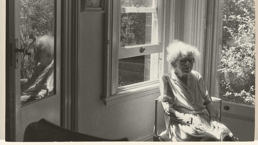 Old black and white photo of Grace Cossington Smith sittng in a sunlit room. She is smiling.