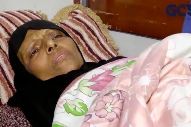 A gravely ill woman lies in a bed under a floral blanket.