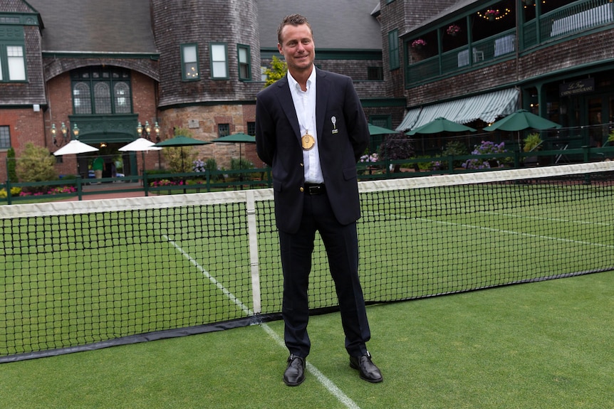 L'Australien Lleyton Hewitt sourit et se tient devant un filet de tennis sur un terrain de gazon portant une médaille d'or autour du cou.