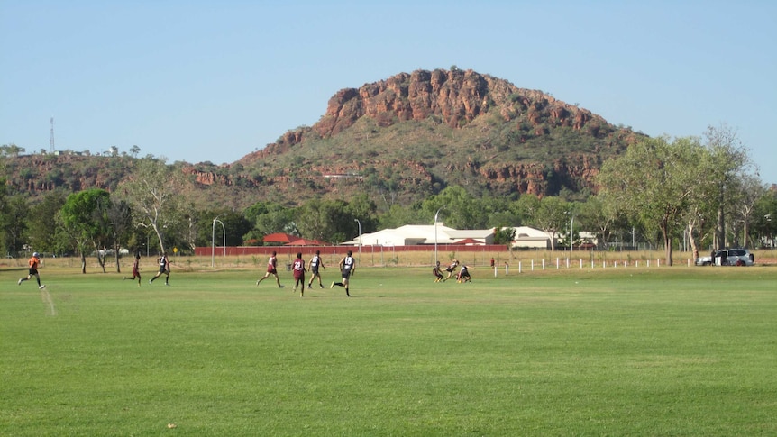 Footballers run across a green oval with a red hill behind
