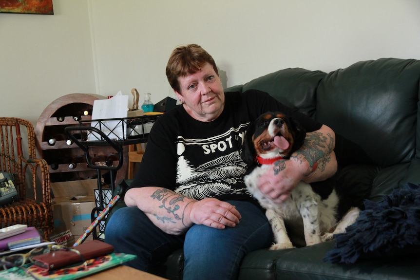 A woman sits on the couch, cuddling her small dog. There are many items around her. Her hair is cropped short.