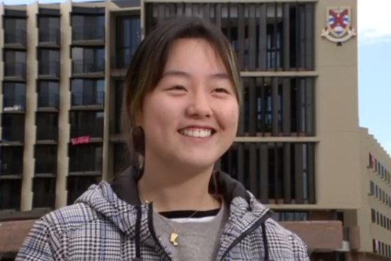A young woman of asian appearance stands in front of a university residential college, smiling.