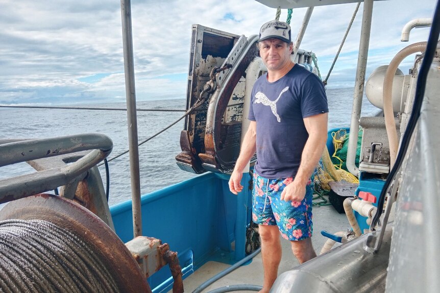 A man in a blue t-shirt and board shorts looks concerned as he stand on the back of a trawler at sea.