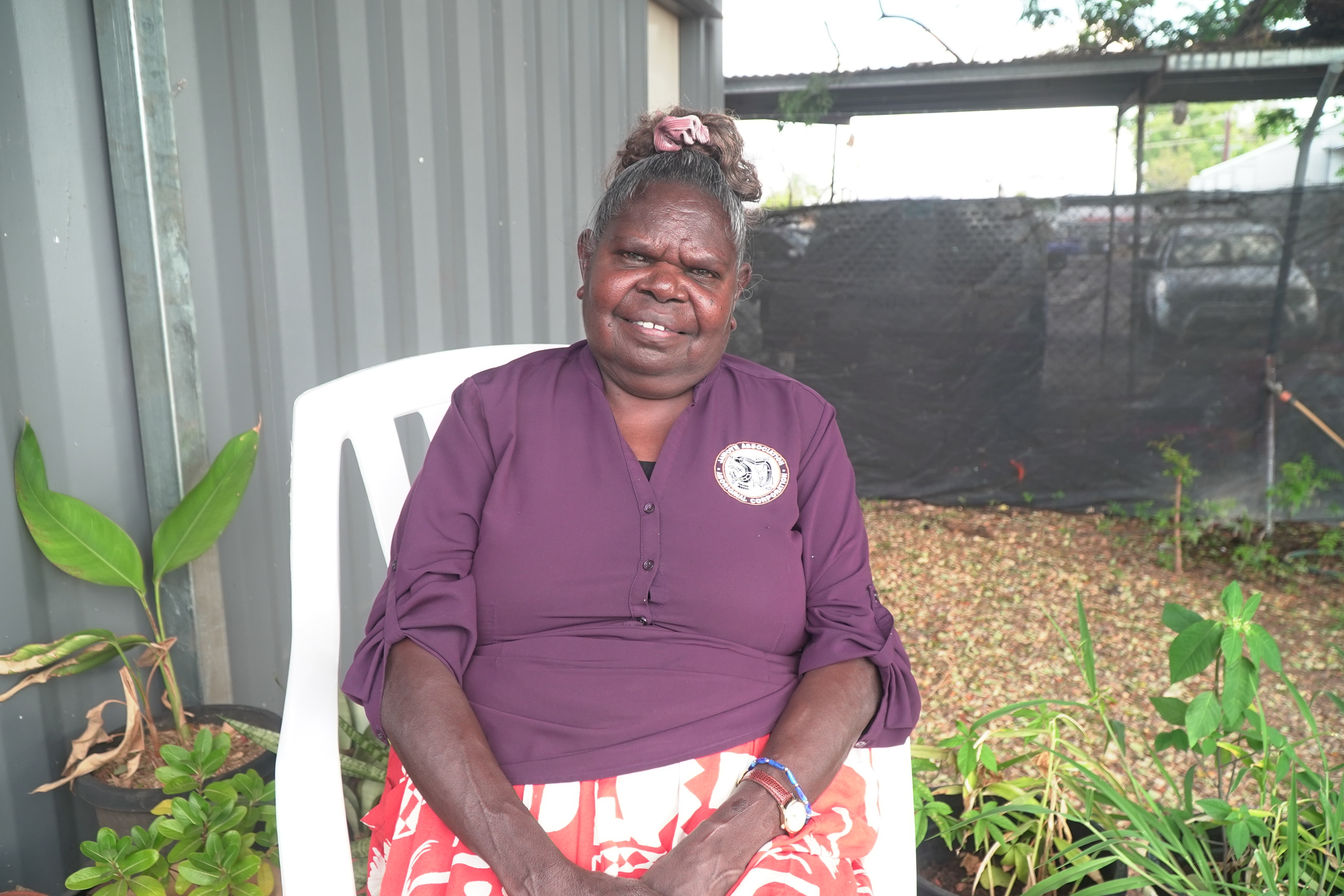 A woman in a purple shirt sits on a plastic white chair in front of a tin shed in a garden