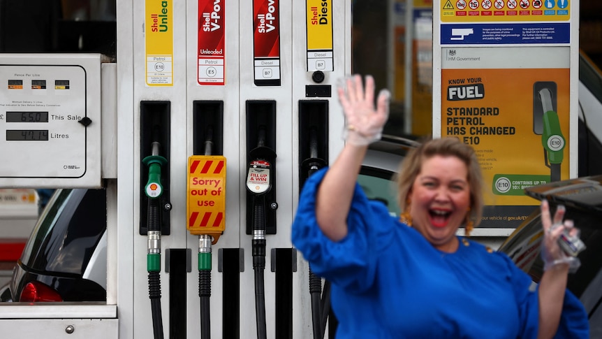 A woman smiles and waves while filling up her car at a petrol station