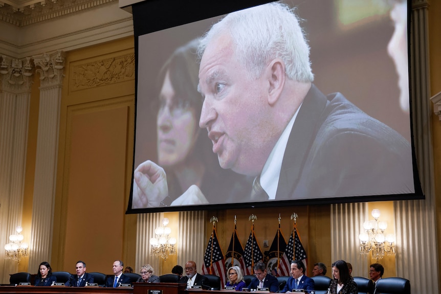 A man with white hair wearing a suit is displayed on a screen above a group of people.