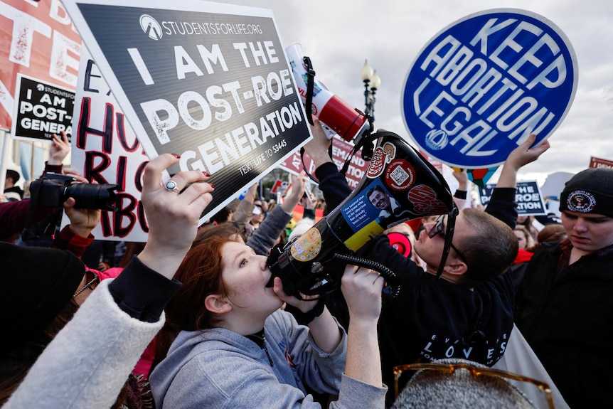 A crowd of protesters with signs about abortion move towards each other, one shouts into a megaphone