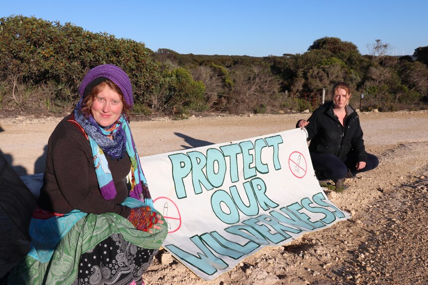Two women sit in the sand holding a sign between them that reads "protect our wilderness"