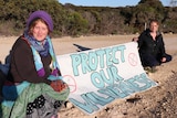 Two women sit in the sand holding a sign between them that reads "protect our wilderness"