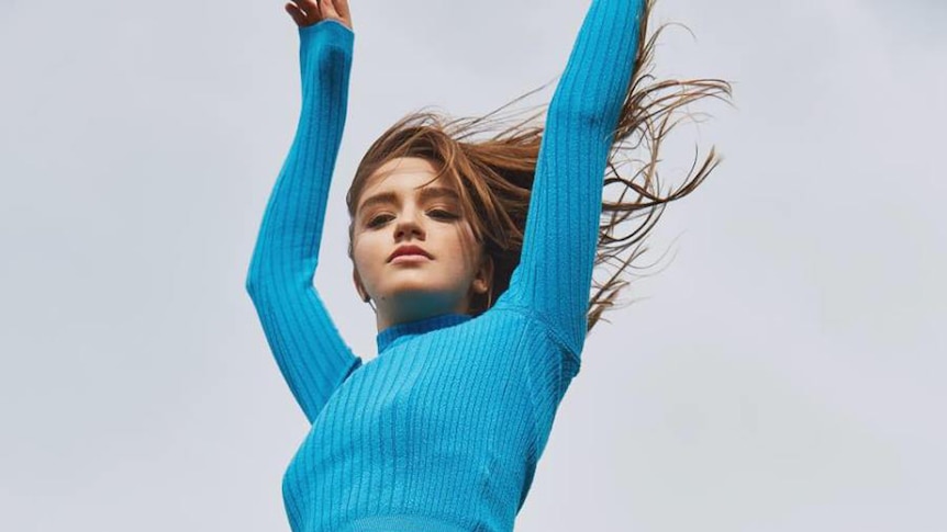 Eves Karydas wears a blue turtleneck and raises her arms to the sky