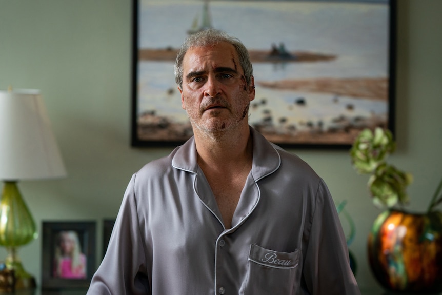 A film still of a pyjama-clad Joaquin Phoenix looking distressed. He has wounds to his head and is standing in a living room.