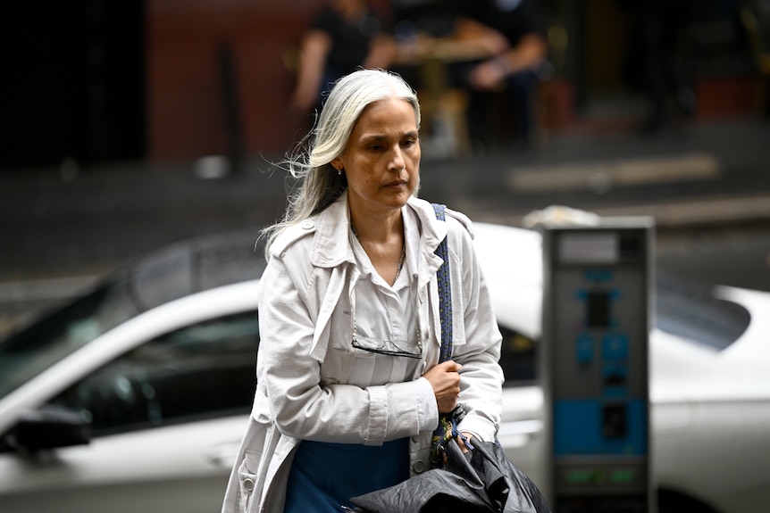 A woman with grey hair and a wait jacket looking somber walks along a street with a silver car in the background