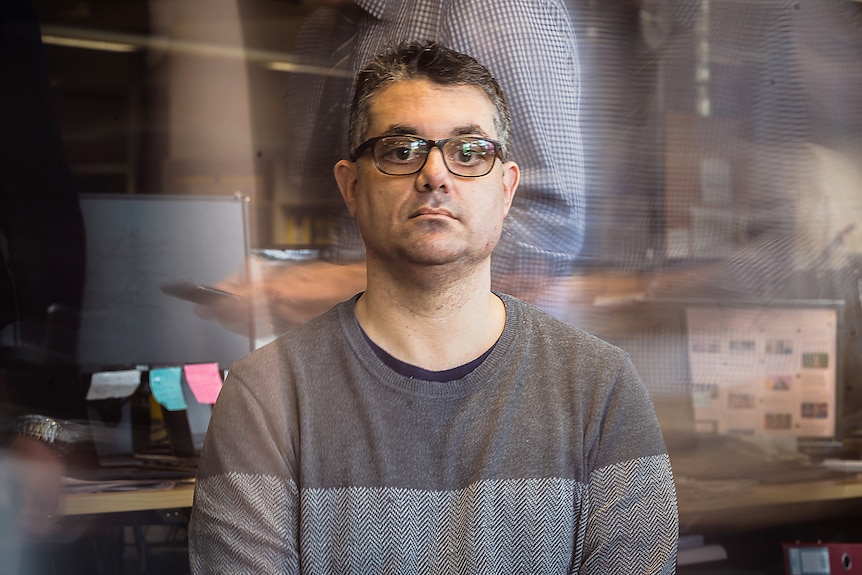 A man with glasses and a grey jumper sits in an office surrounded by blurred figures of office workers.