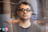A man with glasses and a grey jumper sits in an office surrounded by blurred figures of office workers.