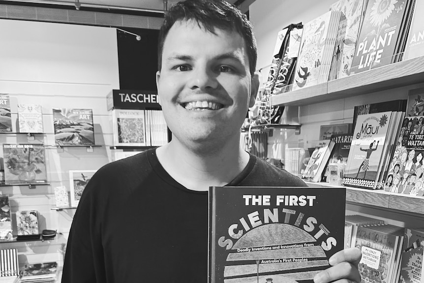 A black and white photo of Corey Tutt who is standing in a bookshop and holding up a copy of his book The First Scientists
